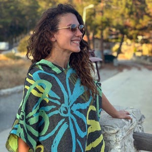 May include: A hooded poncho featuring a dark gray base with large, bold floral patterns in bright turquoise and lime green. The garment has a loose, relaxed fit with short sleeves and a hood, showcasing a textured fabric with a visible weave.