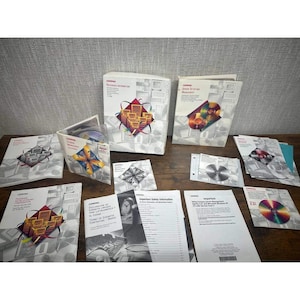 May include: A collection of Compaq technical manuals, documentation booklets, and software discs. The white paper materials feature red and yellow graphic icons, technical text, and diagrams. Several discs are visible, including one with a rainbow-colored surface, all arranged on a wooden surface.
