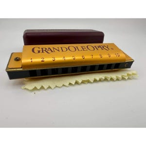 May include: A gold-colored harmonica with the text "GRAND OLE OPRY" printed on the top. The harmonica has black vents and a row of numbered holes. A maroon case rests behind the harmonica, and a yellow cleaning cloth is in front.