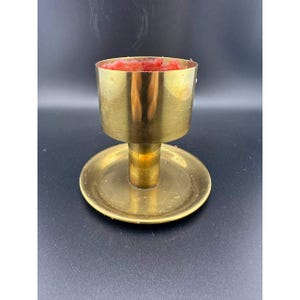 May include: A vintage brass candle holder with a round base and a cylindrical cup. The cup is filled with red wax. The candle holder has a classic design and is likely made of solid brass.