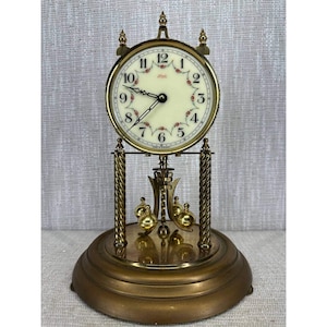 May include: A brass-toned mantel clock with a cream-colored face and floral accents. The clock features a pendulum and a round base. The clock's face has black numerals and hands. The clock is a decorative item.