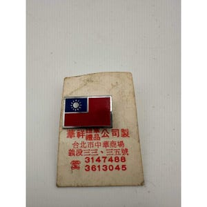 May include: A rectangular pin featuring the flag of Taiwan, with a blue canton and a red field. The pin is set on a worn, cream-colored card with Chinese text and phone numbers.