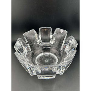 May include: Clear glass bowl with a unique, flower-like design. The bowl has a circular base and a series of outward-facing, rectangular petals. The glass is transparent, allowing light to pass through, and the bowl sits on a dark surface.