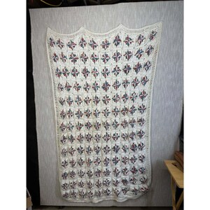 May include: Cream-colored, handmade crochet blanket with a repeating pattern of red and blue star-like shapes. The blanket has a scalloped edge and a textured appearance, suitable for a bed or sofa.
