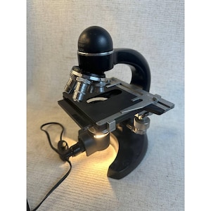 May include: A vintage black microscope with silver-colored metal components. The microscope has a curved arm, a round eyepiece, and a stage for slides. A light source is attached to the base, and a power cord is visible.