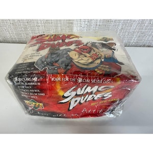 May include: Sealed box of "Sumo Dudes" trading cards. The packaging is red and white with cartoon illustrations of sumo wrestlers. Text includes "Hot New Game" and "Collect all 54 Sumo Dudes discs."