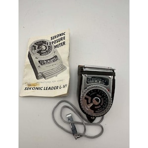 May include: A vintage Sekonic Leader L-VI exposure meter with a silver and black design, accompanied by its instruction manual. The meter has a circular dial and a leather case. A silver chain is attached to the meter.