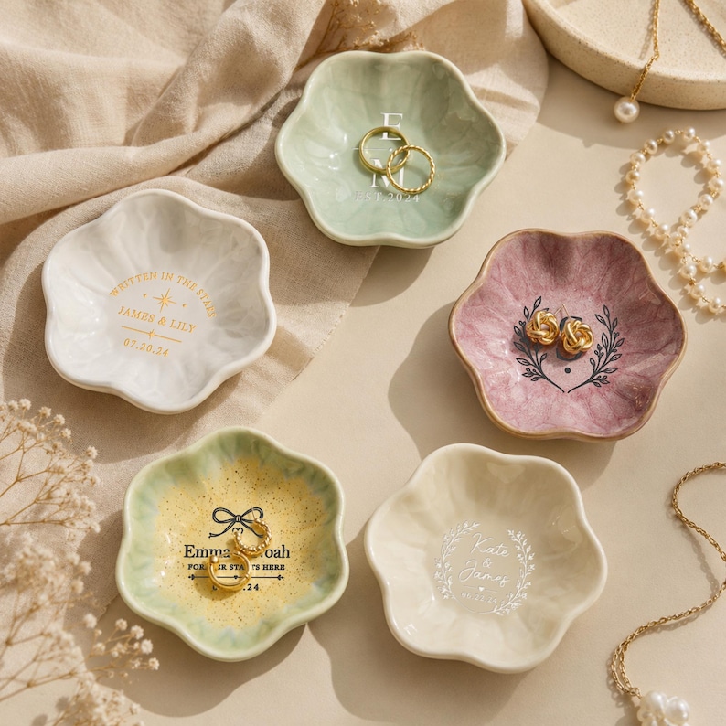 May include: Five flower-shaped ceramic trinket dishes in white, sage green, mauve, and pale yellow. Each dish features unique custom text and graphics, including names, dates, and botanical motifs. Gold-toned rings and earrings are placed inside.