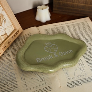 May include: A sage green ceramic tray with an irregular shape. The tray features the text "THE Brook & Gavin" in a silver color, along with a heart-shaped graphic. The tray is resting on a vintage newspaper.