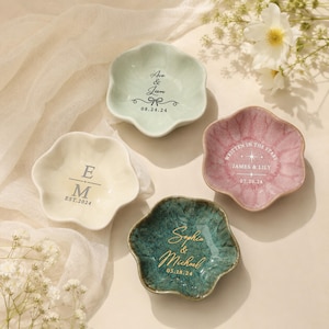Könnte beinhalten: Vier blumenförmige Schmuckschalen aus Keramik in Hellgrün, Creme, Rosa und Dunkel-Türkis. Die Schalen sind mit personalisierten Texten wie Namen, Daten und dekorativen Motiven bedruckt, die jeweils mittig auf den Schalen platziert sind.
