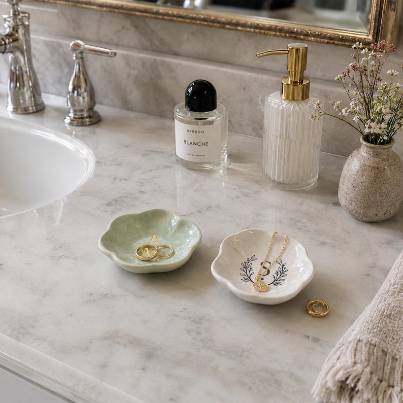 May include: Two small, flower-shaped ceramic trinket dishes sit on a marble countertop. One dish is light green and holds gold rings, while the other is white with a black botanical wreath design and the letter S, holding a gold chain necklace.