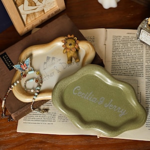 May include: Two decorative ceramic trays. One is beige with a mountain and text design, and a small lion figurine. The other is olive green with the text "Cecilia & Jerry". A beaded necklace and a butterfly brooch are also visible.