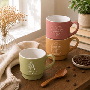 May include: Three ceramic mugs in sage green, mustard yellow, and dusty rose. The green mug features the name Amelia with a floral letter A. The yellow mug displays The Wilson Cafe text. The rose mug shows the name Charlotte inside a heart outline.