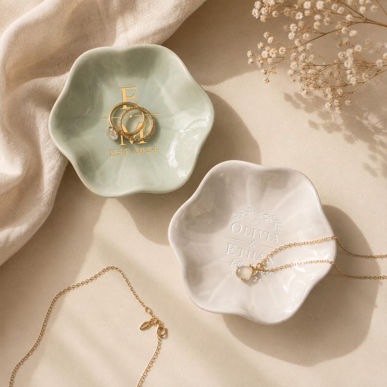 May include: Two flower-shaped ceramic trinket dishes. The sage green dish features gold text reading E M EST. 2024 and holds two gold rings. The white dish features white embossed text reading OLIVIA & ETHAN and holds a gold necklace with a clear stone.