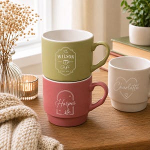 Puede incluir: Una pila de dos tazas de cerámica en verde oliva y rosa empolvado, con una taza blanca al lado. La taza verde presenta el texto The WILSON Café EST. 2026. La taza rosa muestra el nombre Harper con un diseño floral y la blanca dice Charlotte.
