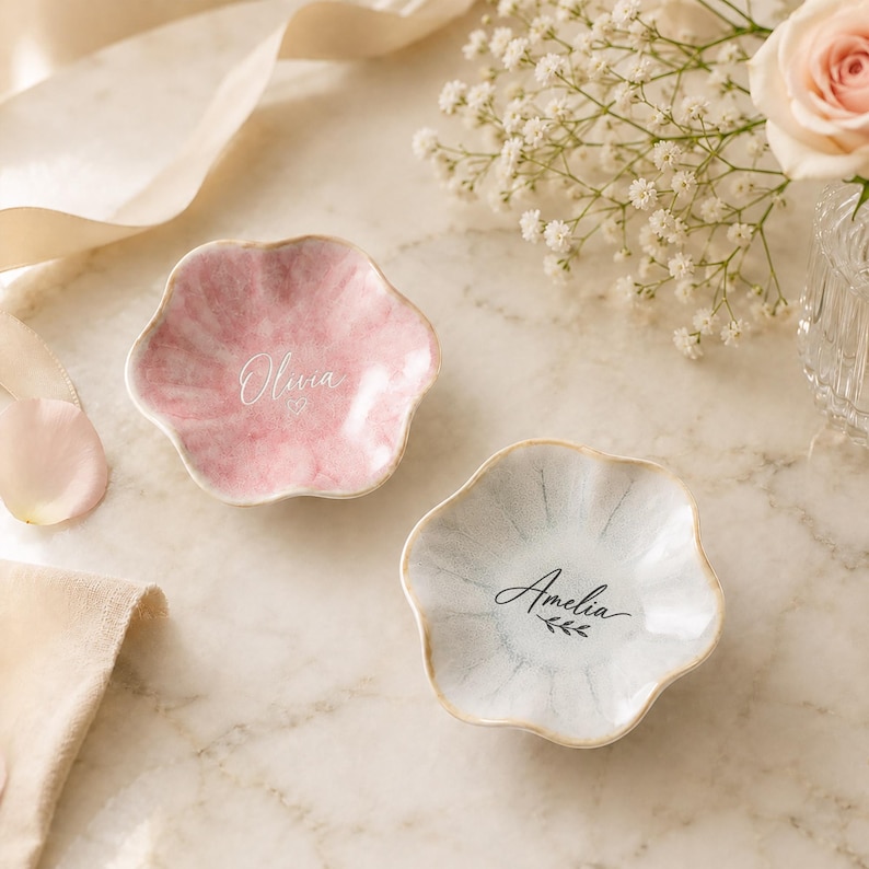May include: Two flower-shaped trinket dishes with scalloped edges. One dish is pink with the name Olivia and a small heart printed in white. The other dish is white with grey accents and the name Amelia printed in black with a leaf icon.