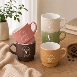 Puede incluir: Un juego de cinco tazas de cerámica en varios colores, incluyendo rosa empolvado, marrón oscuro, beige, verde oliva y amarillo mostaza. Cada taza presenta texto y gráficos impresos en blanco, con nombres como Harper, James y Emma y diseños de café.