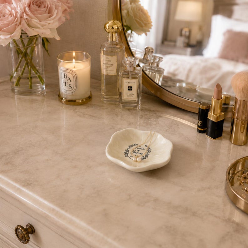 May include: A small, white, scalloped ceramic trinket dish featuring a central laurel wreath design with the text "Kate & James" printed in black. The dish holds a delicate gold chain necklace with a small pearl pendant on a marble surface.