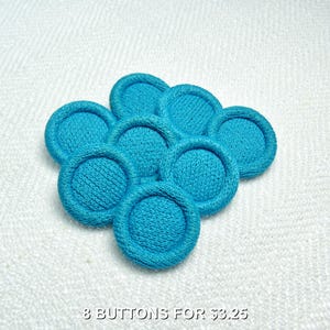 May include: Eight turquoise fabric-covered buttons arranged in a circular pattern. Each button has a textured center and a raised, stitched edge. The buttons are a vibrant shade of blue, and the image is taken on a white surface.