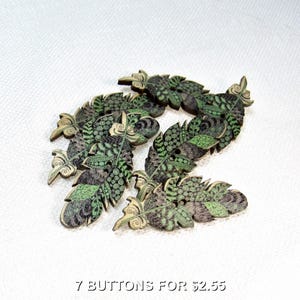 Peut inclure: Sept boutons en bois en forme de plumes. Les boutons présentent un motif détaillé avec des nuances de vert, de marron et de gris. Chaque bouton comporte deux trous pour la couture. Le texte en bas indique "7 BUTTONS FOR $2.55".
