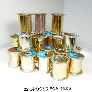 May include: A collection of metallic sewing thread spools arranged in a pyramid formation. Each spool features a cylindrical body with a top label displaying the text Dual Duty and a red bird graphic. The spools have a shiny gold or silver finish.