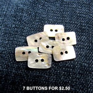 May include: A collection of seven rectangular buttons crafted from iridescent mother-of-pearl material. Each button features two centered holes for attachment and displays a unique, shimmering surface with natural cream and tan tones.
