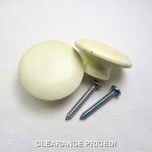 May include: Two round, off-white wooden cabinet knobs with a smooth finish and a slightly domed top. The set includes two silver-colored metal screws with threaded shanks for installation. The text CLEARANCE PRICED! is visible at the bottom.