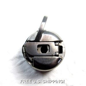 May include: A silver-colored sewing machine bobbin case. The case has a circular shape with a rectangular opening and a curved handle. The text "FREE U.S. SHIPPING!" is at the bottom of the image.