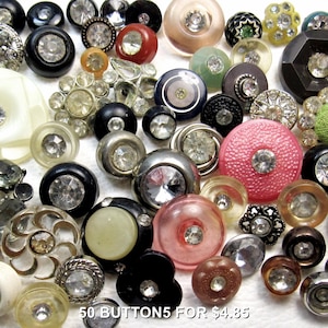 May include: A collection of assorted round buttons featuring embedded clear rhinestones in various colors including black, pink, red, silver, and cream. The buttons vary in size and texture, with some displaying metallic or plastic finishes. Text reads 50 BUTTONS 5 FOR $4.85.