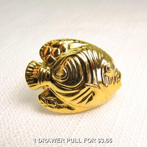 May include: A shiny gold-toned metal drawer pull shaped like a fish. The hardware features embossed details representing scales, fins, and a circular eye. The surface is highly reflective with a smooth, polished finish, capturing light across its curved form.