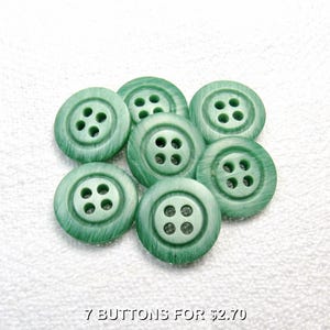 May include: Seven green buttons with four holes each, arranged in a circular pattern. The buttons have a textured appearance and a slightly marbled effect. The text at the bottom reads "7 BUTTONS FOR $2.70".