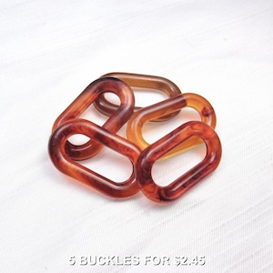 May include: Five oval-shaped buckles in shades of brown and amber are arranged on a white surface. The buckles have a marbled appearance, with darker streaks within the translucent material. The text "5 BUCKLES FOR $2.45" is at the bottom.