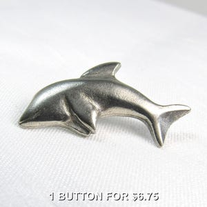 May include: A silver-colored dolphin-shaped button. The button is a detailed representation of a dolphin, with a fin, body, and tail. The text "1 BUTTON FOR $6.75" is at the bottom of the image.