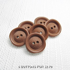 Puede incluir: Un conjunto de seis botones redondos de color marrón, cada uno con un borde exterior liso y elevado y un centro texturizado con dos agujeros. Los botones están dispuestos en grupo, mostrando un color marrón constante y forma circular.