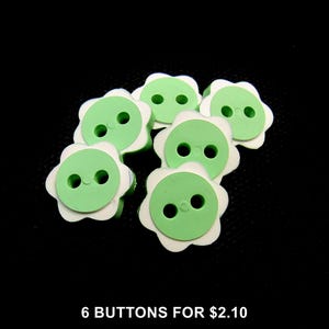 Può includere: Un set di sei bottoni a forma di fiore, ognuno caratterizzato da un centro circolare verde chiaro con due fori e un bordo smerlato bianco. I bottoni sono disposti a grappolo, mettendo in risalto il design floreale e i colori.