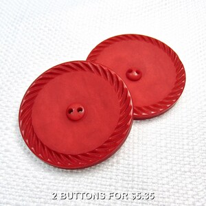 May include: Two round, bright red buttons featuring a central two-hole design and a decorative twisted rope-style border around the outer edge. The buttons have a smooth, matte finish and are positioned overlapping each other on a white textured surface.