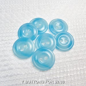 May include: Seven light blue buttons with two holes each, arranged on a white textured surface. The buttons have a marbled appearance, with a lighter blue swirling pattern. The text "7 BUTTONS FOR $2.30" is at the bottom.