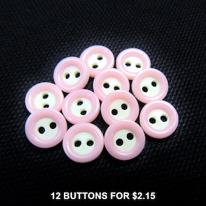 May include: A collection of twelve pink and white buttons with two holes each. The buttons are arranged on a dark textured surface. The buttons have a glossy finish and a classic design, suitable for various sewing projects.