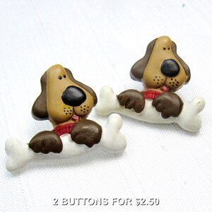 May include: Two buttons shaped like brown and tan dogs with black noses and red collars, each resting their front paws on a white bone. The buttons feature a smooth finish and detailed facial features, including small black dots on the muzzle.
