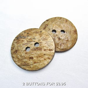 May include: Two round, flat buttons with a textured, mottled brown and tan surface. Each button features two circular holes in the centre for attachment. The buttons have a natural, fibrous appearance with visible grain patterns across the surface.