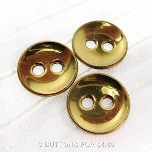 Può includere: Tre bottoni lucidi color oro con due fori ciascuno. I bottoni sono rotondi e hanno una forma leggermente bombata. I bottoni sono su uno sfondo bianco.