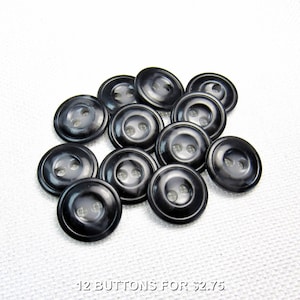 Può includere: Un gruppo di dodici bottoni neri lucidi, ciascuno con due fori. I bottoni sono rotondi con un centro leggermente rialzato e un bordo sottile e scanalato. Il testo "12 BUTTONS FOR $2.75" è in basso.