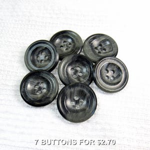 May include: Seven dark grey buttons with a marbled effect. Each button has four holes for sewing. The buttons are arranged on a white textured surface.