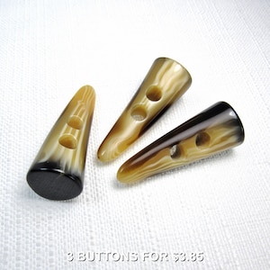 May include: Three horn toggle buttons with a light brown and dark brown marbled pattern. Each button has two or three holes for sewing. The buttons are cone-shaped with a flat, dark brown base.