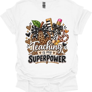 May include: A white t-shirt featuring a graphic design with a leopard print pumpkin, a ruler, a pencil, and scissors. The design includes black bows with white polka dots and the text "Teaching is my SUPERPOWER" surrounded by small leopard print hearts.