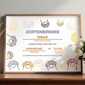 Könnte beinhalten: Eine gerahmte Tieradoptionsurkunde mit dem Namen "Rosalie" und dem Datum "24.07.25". Die Urkunde hat Pfotenabdruck-Illustrationen in verschiedenen Farben und wird auf einer Holzoberfläche mit einer Vase und Blumen präsentiert.