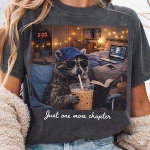 May include: Dark gray t-shirt featuring a raccoon wearing sunglasses and a blue cap, sipping from a drink. The raccoon is in a chair, with a laptop, book, and fairy lights in the background. The text "Just one more chapter" is below the image.