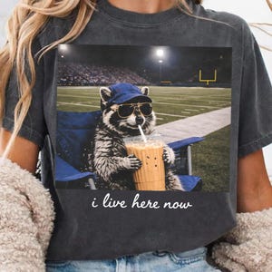 Op de afbeelding: Donkergrijs T-shirt met een wasbeer met een zonnebril en een blauwe pet, zittend in een blauwe stoel, met een drankje met een rietje, met een voetbalveld op de achtergrond. De tekst "i live here now" staat eronder.