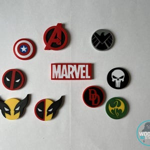 May include: A collection of colorful Marvel character logos and symbols, including Captain America's shield, the Avengers logo, and Deadpool's mask. The word "MARVEL" is displayed in red and white. Various other character emblems are also present.