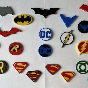 Könnte beinhalten: Eine Sammlung farbenfroher DC Comics Logos und Symbole, darunter Batman, Superman und The Flash. Die Logos haben verschiedene Formen und Farben, wie Gelb, Rot, Schwarz und Blau. Die Artikel sind auf einer weißen Oberfläche angeordnet.